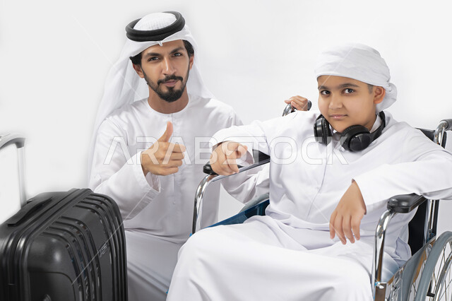 بورتريه لطفل عربي إماراتي خليجي من ذوي الهمم يجلس على كرسي متحرك ويرتدي الزي التقليدي كندورة وعمامه مستعد لسفر مع والده  ، التمكين الاجتماعي  ، اهتمام ودعم المجتمع الإماراتي لذوي  الاحتياجات الخاصه، شعور ذوي الهمم بالراحه والطمأنينه في بيئتهم , خلفية بيضا