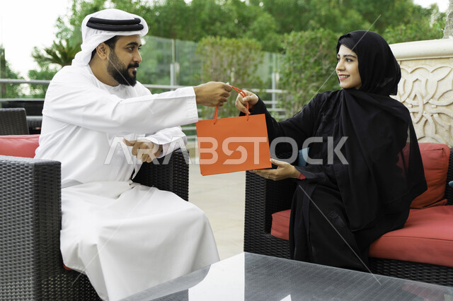 إيماءات السرور والإعجاب بالحصول على هديه في الأعياد والمناسبات السعيده، الاحتفال بمناسبة ذكرى الزواج، زوج عربي اماراتي خليجي يرتدي الكندورة والغترة يقضي اوقات ممتعه برفقه زوجته في أحد المقاهي، سيدة سعودية تجلس في الهواء الطلق مع زوجها.