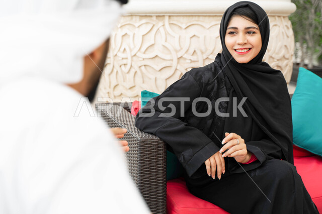 تبادل الدردشات و الأحاديث في الخارج، مفهوم الترابط الفكري و التفاهم بين الزوجين، التنزه في الهواء الطلق، صورة مقربة من الخلف لرجل عربي اماراتي يجلس مع زوجته بأحد المقاهي الفاخرة، سيدة سعودية محجبة ترتدي العباءة الخليجية تقضي أوقات مميزة مع زوجها.