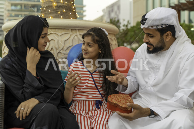  مفهوم التربية بالحب والمودة، تداول الأحاديث و الحوارات بين أفراد الأسرة، بالونات و زينة أعياد الميلاد، زوج عربي اماراتي خليجي يرتدي الكندورة والغترة مع ابنته وزوجته في أحد المقاهي، رجل سعودي يقوم بتقديم هدية على شكل قلب لإبنته، قضاء اوقات عائلية ممتعه.