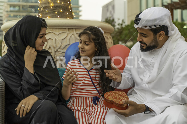 تداول الأحاديث و الحوارات بين أفراد الأسرة، مفهوم التربية بالحب والمودة، بالونات و زينة أعياد الميلاد، زوج عربي اماراتي خليجي يرتدي الكندورة والغترة مع ابنته وزوجته في أحد المقاهي، رجل سعودي يقوم بتقديم هدية على شكل قلب لإبنته، قضاء اوقات عائلية ممتعه.
