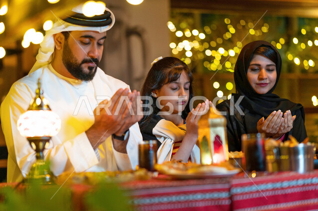 A family Ramadan breakfast in a restaurant decorated with Ramadan, Islamic and worship decorations, a close-up of an Emirati Arab family sitting on the dining table and praying to God at the time of breaking the fast in the blessed month of Ramadan, a Sau