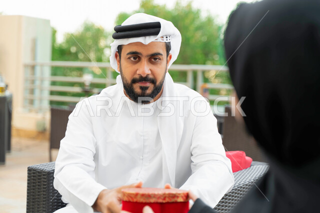 نظرات المحبة والاهتمام بين الزوجين وإحياء ذكرى يوم الزواج, جلسات ممتعه خارج المنزل, رجل عربي خليجي إماراتي يقدم لزوجته هدية في علبة حمراء بمناسبة عيد الحب, زوجان سعوديان يجلسان في الهواء الطلق في إحدى المنتجعات الفاخره,  إيماءات تدل على السعادة والسرور