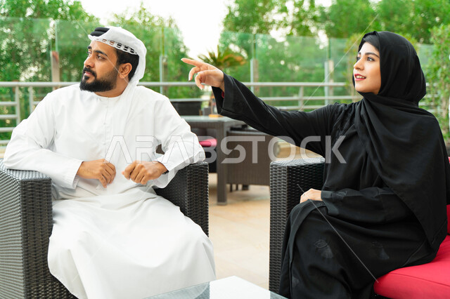 سيدة سعودية محجبة ترتدي العباءة الخليجية و تشير الى شيء ما، زوجان عربيان اماراتيان يقومان بالتنزه في الهواء الطلق بأحد المنتجعات الفاخرة، قضاء أوقات مميزة بين الزوجين و تبادل الاحاديث والدردشات في الخارج، الاشارة الى شيء معين