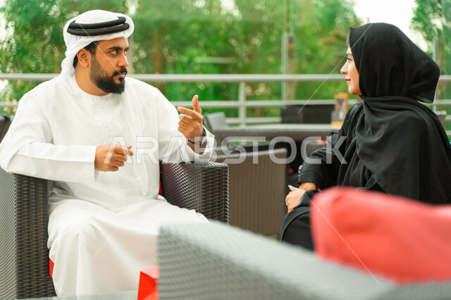 التفاهم والترابط الفكري بين الزوجين، تبادل الدردشات والأحاديث في الخارج، التنزه في الهواء الطلق، رجل عربي اماراتي يجلس مع زوجته بأحد المقاهي الفاخرة، سيدة سعودية محجبة ترتدي العباءة الخليجية تقضي أوقات مميزة مع زوجها، ايماءات يدين تدل على الاندماج بالحديث