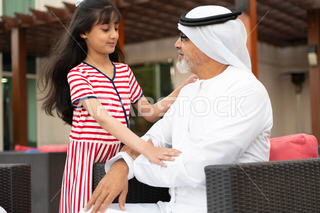 التسامر والضحك مع الأحفاد, إحترام وتقدير الأجداد وكبار السن، الزيارات و التجمعات العائلية، أجواء عائلية ممتعة في الهواء الطلق، ايماءات تدل على السعادة, صورة مقربة لرجل مسن عربي خليجي إماراتي ينظر إلى حفيدته بحب, فتاة سعودية وجدها في جلسة خارجيه راقية .