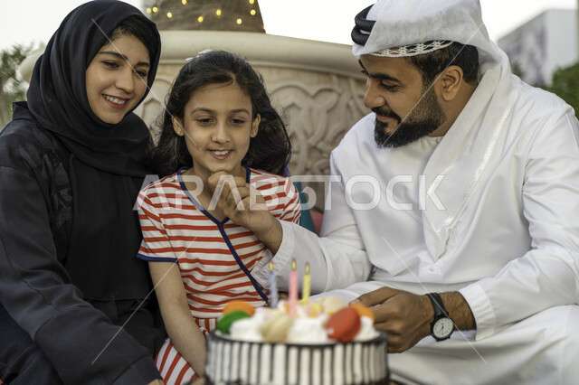 مفهوم الترابط الأسري، عائلة عربية خليجية إماراتية تقضي أوقات ممتعه في الخارج، صورة مقربة لأسرة سعودية تحتفل بعيد ميلاد ابنتها في أحد المقاهي، أجواء اسرية مليئة بالحب والحنان، إيماءات تدل على السعادة باجتماع افراد العائلة, كعكة يوم الميلاد المزينة بالشموع 