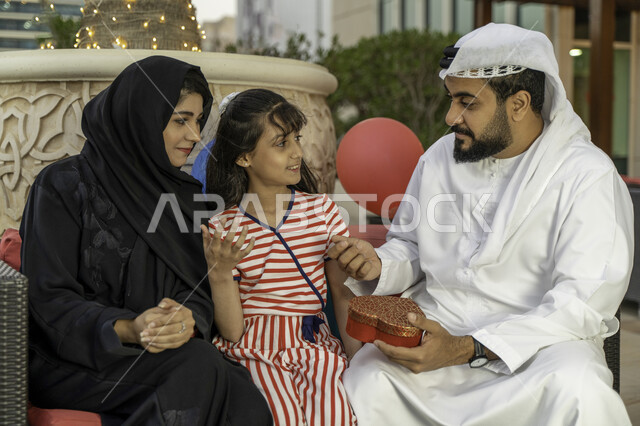 الدردشة وتبادل الأحاديث بين أفراد الأسرة، مفهوم التربية بالحب والمودة، زينة وبالونات أعياد الميلاد، رجل عربي اماراتي خليجي يرتدي الكندورة والغترة مع زوجته و ابنته في أحد المقاهي، الاحتفال بالمناسبات العائلية السعيدة، أب سعودي يقوم بتقديم علبة هدية لإبنته