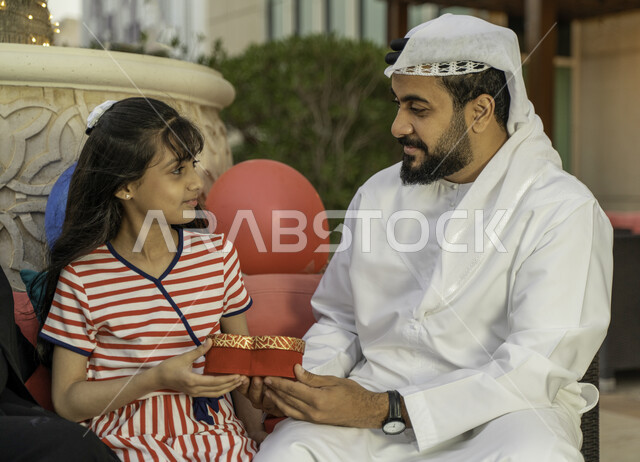  مفهوم التربية بالحب والمودة، بالونات أعياد الميلاد، رجل عربي اماراتي خليجي يرتدي الكندورة والغترة يحتفل مع ابنته بيوم ميلادها في أحد المقاهي، المناسبات العائلية السعيدة، شاب سعودي يقوم بتقديم علبة هدية لإبنته، الاهتمام والاعتناء بالأبناء