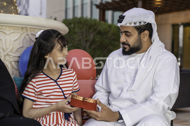 بالونات أعياد الميلاد، مفهوم التربية بالحب والمودة، رجل عربي اماراتي خليجي يرتدي الكندورة والغترة يحتفل مع ابنته بيوم ميلادها في أحد المقاهي، المناسبات العائلية السعيدة، شاب سعودي يقوم بتقديم علبة هدية لإبنته، الاهتمام والاعتناء بالأبناء
