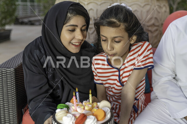 البهجة والفرحة في المناسبات العائلية السعيدة, ايماءات النفخ على الشموع, صورة مقربة لإمرأة عربية خليجية إماراتية تحتفل بعيد ميلاد طفلتها, أم سعودية وابنتها يقومان بإطفاء شموع الميلاد خارج المنزل, احتفال عائلي بسيط في الهواء الطلق
