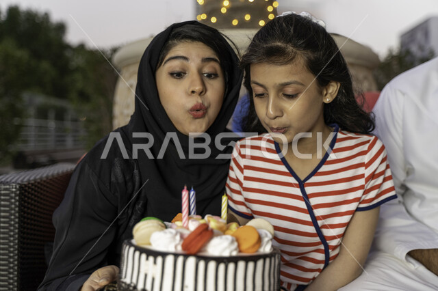 Joy and joy in happy family occasions, gestures blowing on candles, a close-up of an Emirati Arab woman celebrating her child's birthday, a Saudi mother and her daughter extinguishing birthday candles outside the house, a simple family celebration in the 