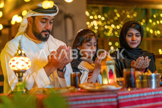 فطور رمضاني عائلي في مطعم مزين بديكورات رمضانية, اسلاميات وعبادة, صورة مقربة لعائلة عربية خليجية إماراتية تجلس على سفرة الطعام وتدعو الله عند موعد الإفطار في شهر رمضان المبارك, أسرة سعودية تشكر الله تعالى بعد تناول طعام الغداء