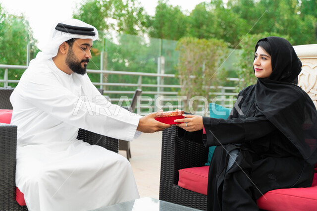 جلسات ممتعه خارج المنزل, رجل عربي خليجي إماراتي يقدم لزوجته هدية في علبة حمراء بمناسبة عيد الحب, زوجان سعوديان يجلسان في الهواء الطلق في إحدى المنتجعات الفاخره, التعبير عن الألفة والمحبة بين الزوجين وإحياء ذكرى يوم الزواج, إيماءات تدل على السعادة والسرور