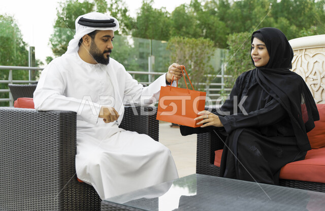 إيماءات الفرح والسعادة, تقديم الهدايا في الأعياد والمناسبات, رجل عربي خليجي إماراتي يقدم لزوجته هديه داخل كيس بمناسبة عيد الأم, زوجان سعوديان يجلسان في الهواء الطلق في إحدى المنتجعات الفاخره, التعبير عن الألفة والمحبة بين الزوجين, إحياء ذكرى يوم الزواج.