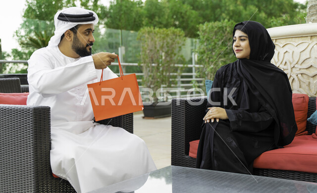 تقديم الهدايا في الأعياد والمناسبات, إيماءات الفرح والسعادة, رجل عربي خليجي إماراتي يقدم لزوجته هديه داخل كيس بمناسبة عيد الأم, زوجان سعوديان يجلسان في الهواء الطلق في إحدى المنتجعات الفاخره, التعبير عن الألفة والمحبة بين الزوجين, إحياء ذكرى يوم الزواج.