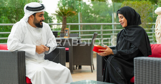 التعبير عن الألفة والمحبة بين الزوجين, جلسات ممتعه خارج المنزل, رجل عربي خليجي إماراتي يقدم لزوجته هديه في علبه حمراء بمناسبة عيد الحب, زوجان سعوديان يجلسان في الهواء الطلق في إحدى المقاهي الفاخره, إحياء ذكرى يوم الزواج, إيماءات السعادة والسرور.