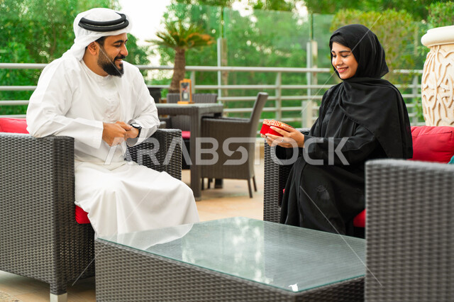Fun sessions outside the home, an Arab Gulf Emirati man presents his wife with a gift in a red box on the occasion of Valentine's Day, a Saudi couple sits outdoors in one of the luxurious cafes, expressing intimacy and love between the spouses, commemorat
