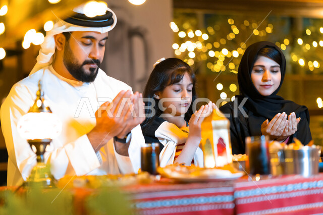 فطور رمضاني عائلي في مطعم مزين بديكورات رمضانية, اسلاميات وعبادة, عائلة عربية خليجية إماراتية تجلس على سفرة الطعام وتدعو الله عند موعد الإفطار في شهر رمضان المبارك, عائلة سعودية تشكر الله تعالى بعد تناول طعام الغداء