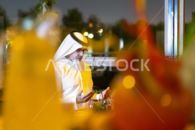 إفطار الصائم عند أذان المغرب, اسلاميات وروحانيات رمضانية, رجل مسن إمارتي عربي خليجي يرتدي الزي الإماراتي يجلس على مائدة الطعام ويدعو الله عند موعد الإفطار في شهر رمضان المبارك, رجل سعودي كبير بالسن يشكر الله بعد تناول طعام الغداء