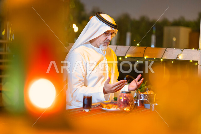 إسلاميات وروحانيات رمضانية, إفطار الصائم عند أذان المغرب, رجل مسن إمارتي عربي خليجي يرتدي الزي الإماراتي يجلس على مائدة الطعام ويدعو الله عند موعد الإفطار في شهر رمضان المبارك, رجل سعودي كبير بالسن يشكر الله بعد تناول طعام الغداء
