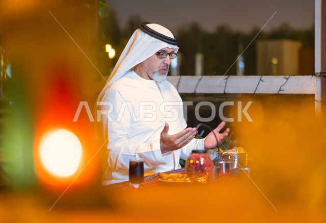 إفطار الصائم عند أذان المغرب, إسلاميات وروحانيات رمضانية, رجل مسن إمارتي عربي خليجي يرتدي الزي الإماراتي يجلس على مائدة الطعام ويدعو الله عند موعد الإفطار في شهر رمضان المبارك, رجل سعودي كبير بالسن يشكر الله بعد تناول طعام الغداء