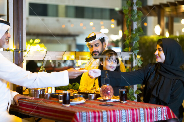 مشاركة الطعام وإكرام الوالدين, سفرة إفطار مليئة بأصناف من المأكولات والمقبلات الشهية, عائلة عربية خليجية إماراتية مستمتعة بتناول الفطور في شهر رمضان, أسره سعودية تجلس في مطعم مزين بديكورات رمضانيه, إجتماع الأهل على مائدة السحور, أجواء عائلية رمضانية.