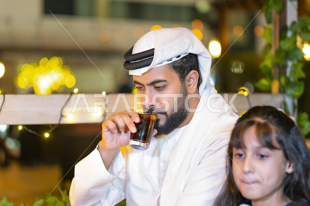 Ramadan spirituality and religious family atmosphere, the holy month of Ramadan, a close-up of an Emirati Arab man with his daughter next to him sitting in a restaurant decorated with Ramadan decorations, a Saudi man enjoying a cold drink with Iftar, brea