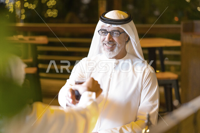 مشاركة المشروبات البارده مع الأهل والأقارب, صورة مقربة لرجلان عربيان خليجيان اماراتيان يرتديان الكندوره والغترة الاماراتيه يجلسان في مقهى, إجتماع رجلا أعمال سعوديان في خارج الشركه, النقاشات والحوارات في الأجواء الهادئة, إكرام وإحترام الوالدين وكبار السن.