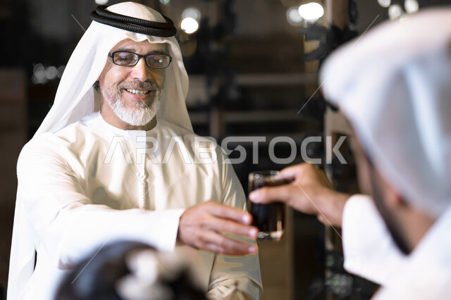 مشاركة المشروبات مع الأهل والأقارب, إكرام وإحترام الوالدين وكبار السن, صورة مقربة لرجلان عربيان خليجيان اماراتيان يرتديان الكندوره والغتره يجلسان في مقهى, إجتماع رجلا أعمال سعوديان خارج الشركه, النقاشات والحوارات في الأجواء الهادئة.