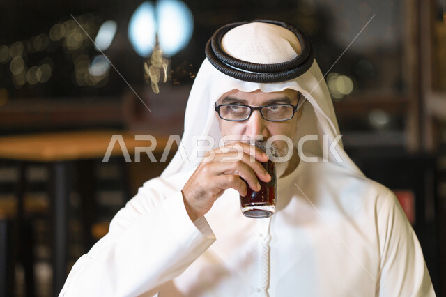 Spending fun times in one of the high-end cafes and restaurants, a close-up of an elderly Arab Emirati man wearing a kandora and an Emirati ghutra, an elderly Saudi man enjoying a cold drink, enjoying the quiet atmosphere outside the house, looking at som