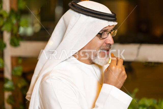 Eating the Ramadan Iftar at the Maghrib call to prayer, a close-up of an elderly Arab Emirati man wearing an Emirati kandura and ghutra spending an enjoyable time alone outside, an elderly Saudi man enjoying himself in one of the high-end cafes and restau