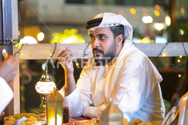 قضاء وقت ممتع في الخارج, النظر إلى شيء ما, كسر الصيام عند أذان المغرب، صورة مقربة لرجل عربي خليجي اماراتي يجلس على مائدة الإفطار في شهر رمضان المبارك, رجل سعودي يتناول الطعام في مطعم مزين بديكورات رمضانية، روحانيات رمضانيه وأجواء دينيه.