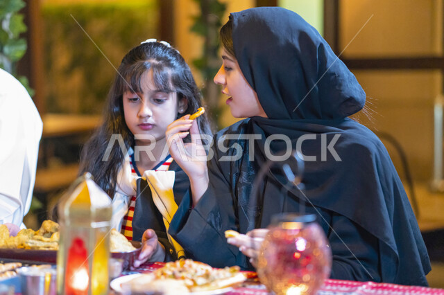 Iftar table filled with varieties of delicious food and appetizers, a close-up of an Arab Emirati woman sitting with her daughter at the breakfast table in the holy month of Ramadan, a Saudi girl with her mother enjoying food, the family gathers on a luxu