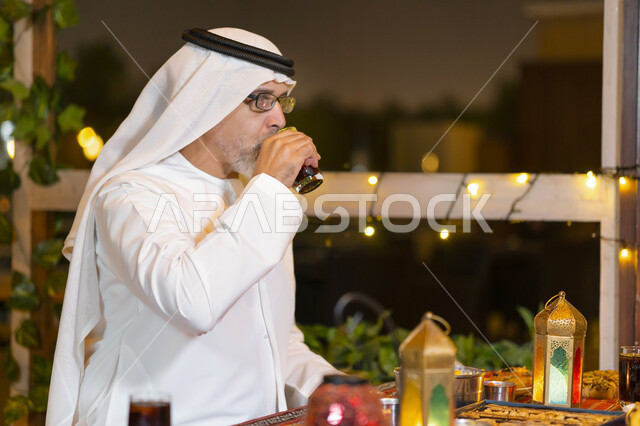 روحانيات رمضانية وأجواء دينية, صورة مقربة لرجل مسن عربي خليجي إماراتي يرتدي الزي الاماراتي التقليدي يجلس في مطعم مزين بديكورات رمضان, فوانيس وزينة رمضانية, رجل سعودي يستمتع بالشراب البارد مع الإفطار, كسر الصيام عند أذان المغرب, شهر رمضان المبارك.