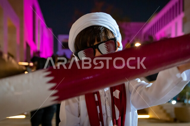 صورة مقربة لطفل عربي خليجي قطري يمسك بوشاح قطر، علم دولة قطر، تشجيع المنتخب الوطني القطري، كأس العالم في قطر 2022، بطولة كأس العالم، فيفا 2022، مونديال قطر، السياحة في قطر