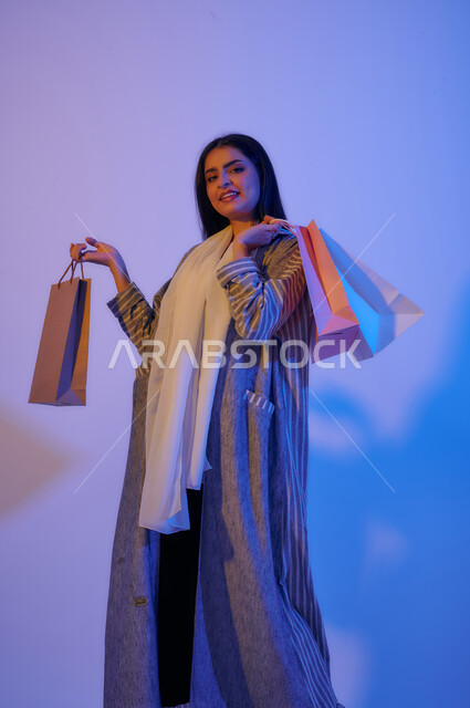 Portrait of a Saudi Arabian Gulf woman wearing an elegant colorful abaya carrying shopping bags in her hand, electronic shopping, international brands, shopping in malls, shopping pleasure in Saudi Arabia, colored shopping bags, seasonal offers and discou