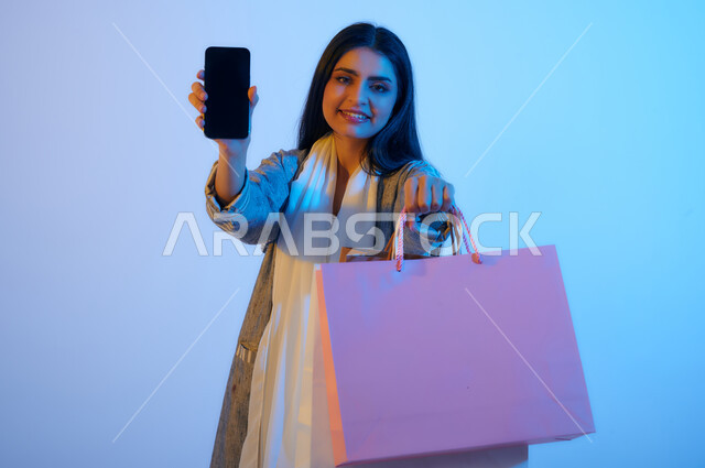 بورتريه لإمرأة عربيه خليجية سعوديه تحمل بيدها أكياس التسوق وجهاز الهاتف المحمول, عرض شاشه سوداء, التسوق الإلكتروني, تطبيقات الهاتف المحمول, ماركات عالمية, متعة التسوق في السعودية, حقائب ورقية, عروض وتخفيضات موسميه 