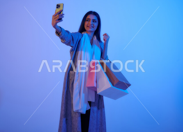 Portrait of a Saudi Arabian Gulf woman wearing a colorful cloak, carrying shopping bags in her hand, taking souvenir photos via mobile phone, online shopping, international brands, shopping in malls, the fun of shopping in Saudi Arabia, colorful shopping 