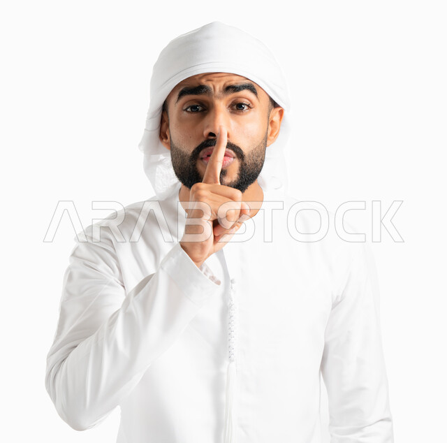 التوقف عن الكلام وخفض الصوت، بورتريه لرجل عربي خليجي إماراتي يرتدي العمامة والكندورة الإماراتية ويضع أصبع السبابة على فمه، الهدوء والسكون وعدم الإزعاج، إيماءات وجه ويدين تدل على طلب السكوت والصمت، حركة أصبع وإشارات، خلفية بيضاء