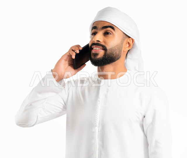 دردشة ومحادثات وتعارف، استخدام الهاتف المحمول في الاتصال والتواصل مع الأفراد، بورتريه لرجل عربي خليجي إماراتي يرتدي العمامة والكندورة الإماراتية ويجري مكالمة هاتفية من خلال الجوال، خلفية بيضاء