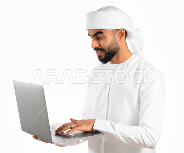 إحتراف وظائف الحاسوب وتكنولوجيا المعلومات، بورتريه لرجل عربي خليجي إماراتي يرتدي العمامة والكندورة الإماراتية يحمل بيده جهاز الحاسوب المحمول، تصفح الإنترنت والعمل عن بعد، إنجاز مهام العمل من خلال اللابتوب، الضغط على أزرار لوحة المفاتيح، خلفية بيضاء