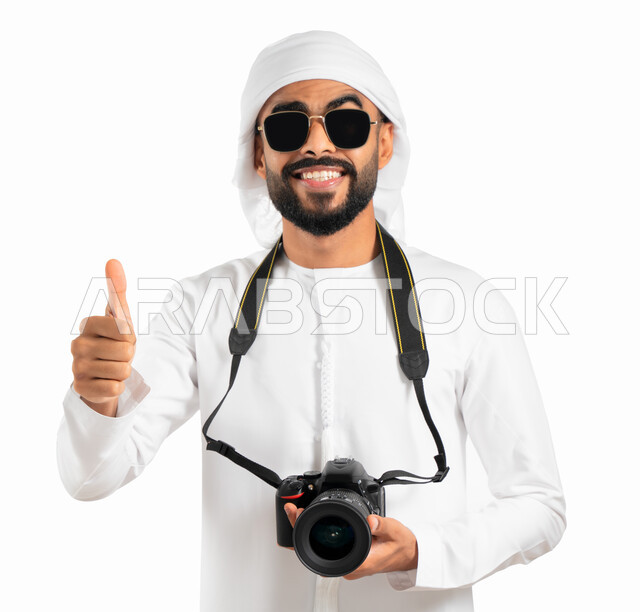 رفع أصبع الإبهام لأعلى بإشارة القبول والرضى، بورتريه لمصور عربي إماراتي خليجي يرتدي الكندورة والعمامة الإماراتية والنظارات الشمسية، إيماءات وجه ويدين تدل على السعادة والإعجاب، إلتقاط الصور الفوتوغرافية بإستخدام كاميرا التصوير عالية الدقة، خلفية بيضاء