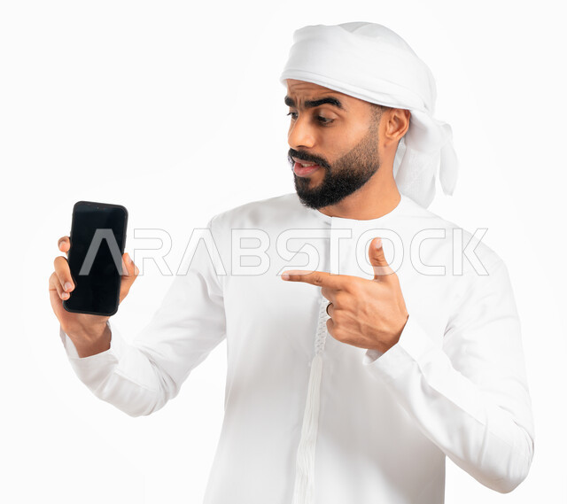 Using smartphone software, a portrait of an Arab Gulf Emirati man wearing a kandora and an Emirati turban and pointing his index finger at a mobile phone with a blank black screen, e-marketing and displaying mobile services and applications, white backgro
