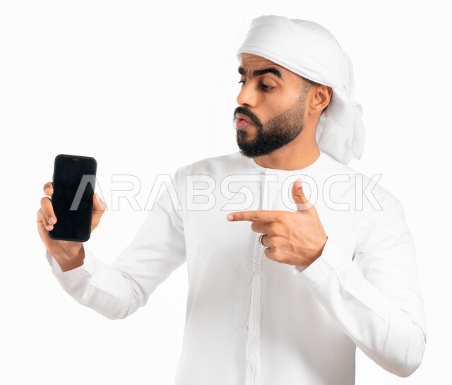 بورتريه لرجل عربي خليجي إماراتي يرتدي الكندورة والعمامة الإماراتية ويشير بإصبع السبابة نحو الهاتف المحمول بشاشة سوداء فارغة، التسويق الإلكتروني وعرض خدمات وتطبيقات الجوال، إستخدام برامج الهاتف الذكي، خلفية بيضاء 