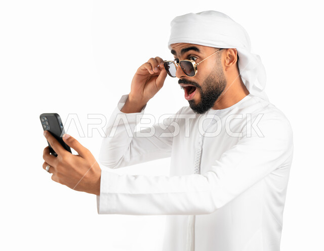 تصفح تطبيقات وبرامج الجوال، بورتريه لرجل عربي خليجي إماراتي يرتدي الكندورة والعمامة الإماراتية وينظر إلى الهاتف المحمول بدهشة وذهول وإنبهار، إرتداء النظارات الشمسية، إيماءات وجه تدل على التعجب والإستغراب، إستخدام وسائل التواصل الإجتماعي، خلفية بيضاء