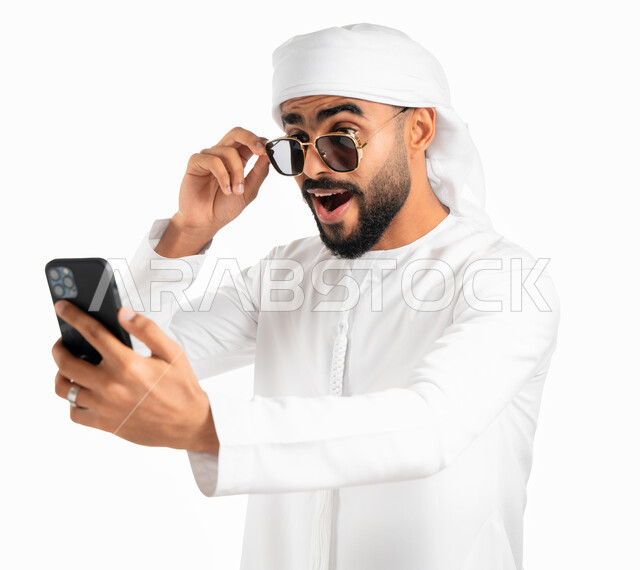 إرتداء النظارات الشمسية، إيماءات وجه تدل على الدهشة وذهول وإنبهار، بورتريه لرجل عربي خليجي إماراتي يرتدي الكندورة والعمامة الإماراتية وينظر إلى الهاتف المحمول بتعجب وإستغراب، إستخدام وسائل التواصل الإجتماعي، تصفح تطبيقات وبرامج الجوال، خلفية بيضاء