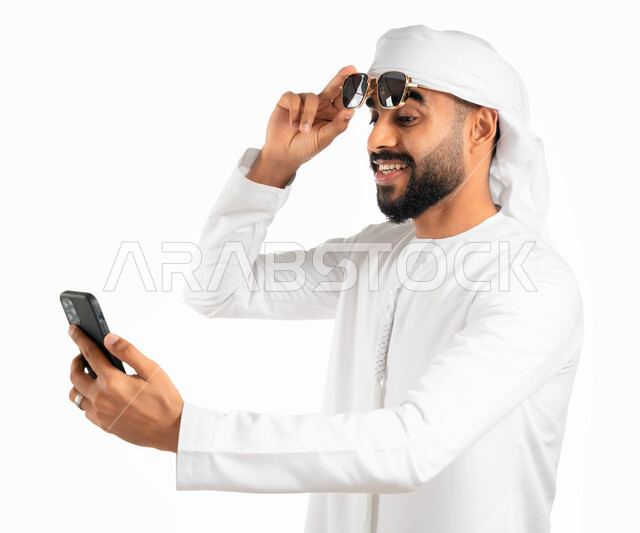 النظر إلى الهاتف المحمول بدهشة وذهول وإنبهار، بورتريه لرجل عربي خليجي إماراتي يرتدي الكندورة والعمامة الإماراتية ويقوم برفع النظارات الشمسية عن عينه، إيماءات وجه تدل على التعجب والإستغراب، إستخدام وسائل التواصل الإجتماعي، إجراء مكالمة فيديو، خلفية بيضاء