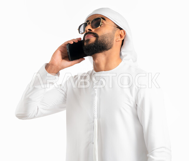 اجراء مكالمة هاتفية من خلال الجوال، بورتريه لرجل عربي خليجي إماراتي يرتدي العمامة والكندورة الإماراتية والنظارات الشمسية، استخدام الهاتف المحمول في الاتصال والتواصل مع الأفراد، دردشة ومحادثات وتعارف، خلفية بيضاء