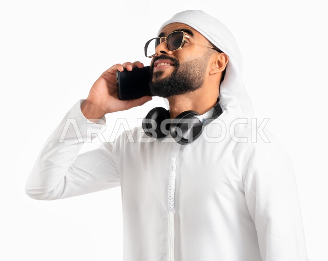 ارتداء النظارات الشمسية ووضع سماعة الرأس حوال الرقبة، بورتريه لرجل عربي خليجي إماراتي يرتدي العمامة والكندورة الإماراتية ويجري مكالمة هاتفية من خلال الجوال، استخدام الهاتف المحمول في الاتصال والتواصل مع الأفراد، دردشة ومحادثات وتعارف، خلفية بيضاء